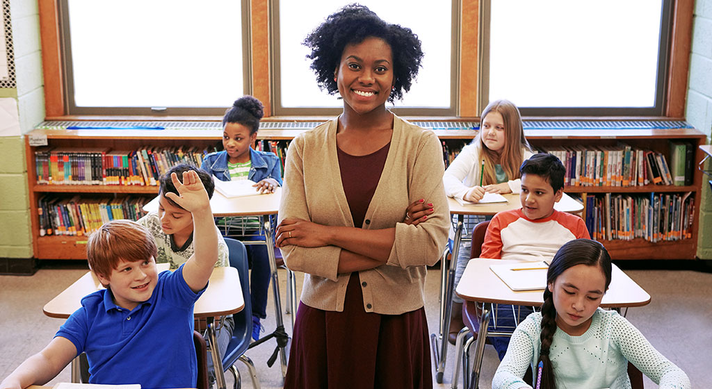 women standing with her arms crossed and smiling in a classroom with young students.